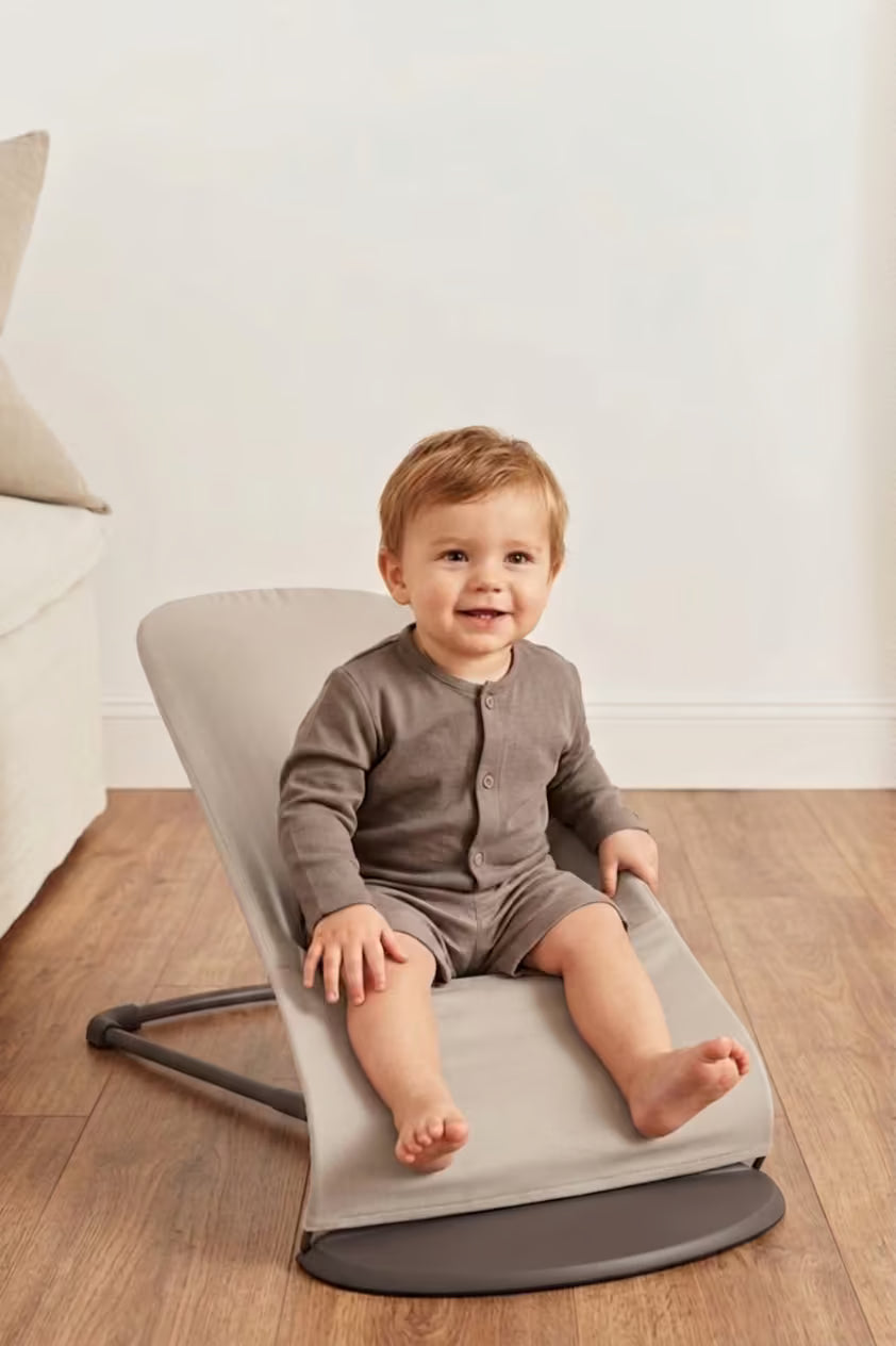 Child sitting on a gray baby bouncer in a room with wooden flooring and a white wall.