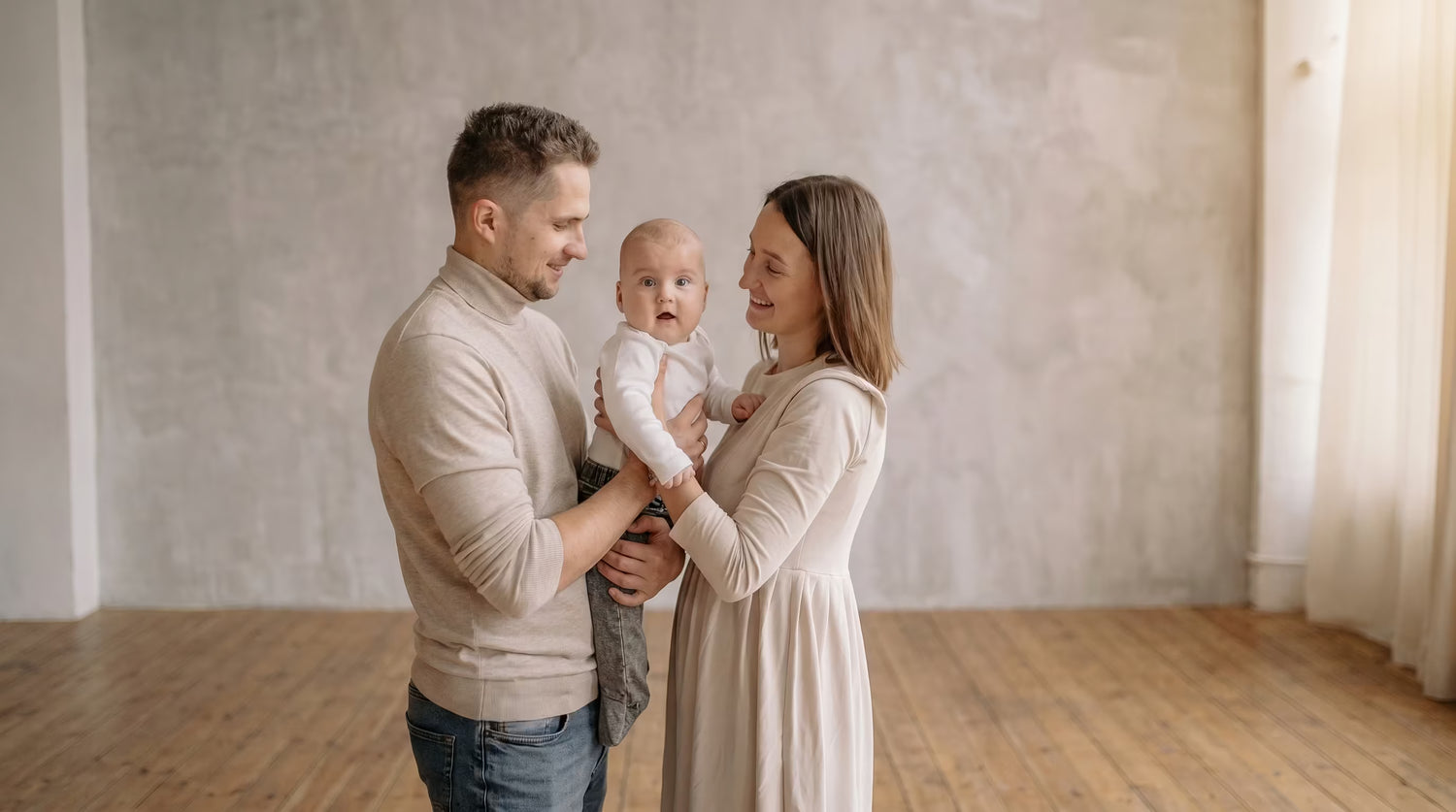 Family of three standing in a room with wooden flooring and white walls.
