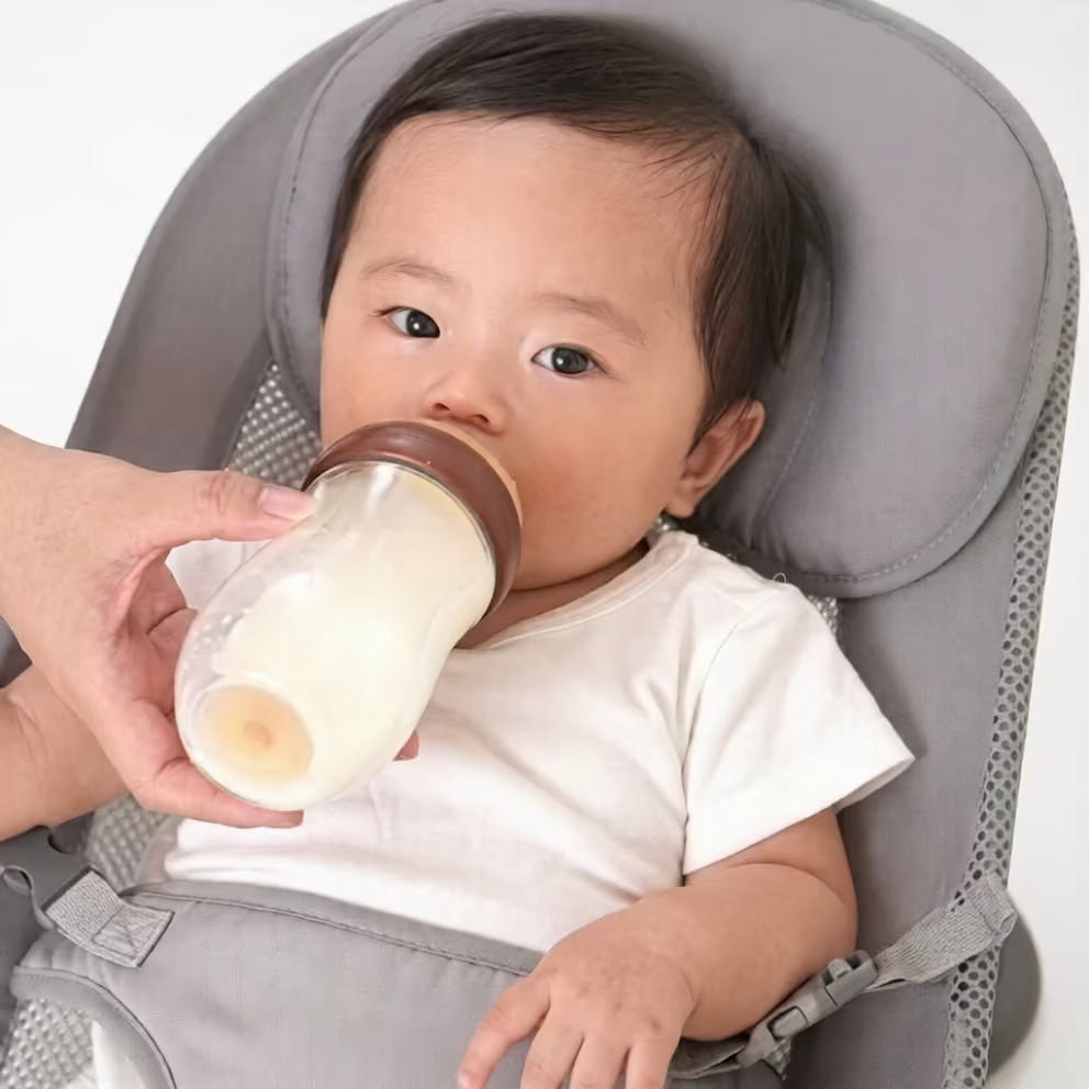 Baby in a high chair being fed from a bottle with a white background.