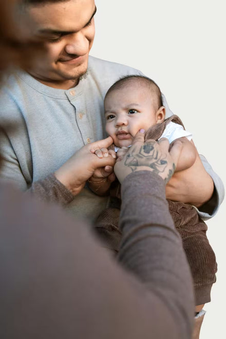 Man holding a baby with another person's hand on the baby's foot against a plain background